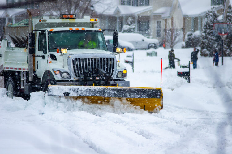 Front view of city services snow plow truck with yellow blade cleaning roads after winter storm with kids playing in the background