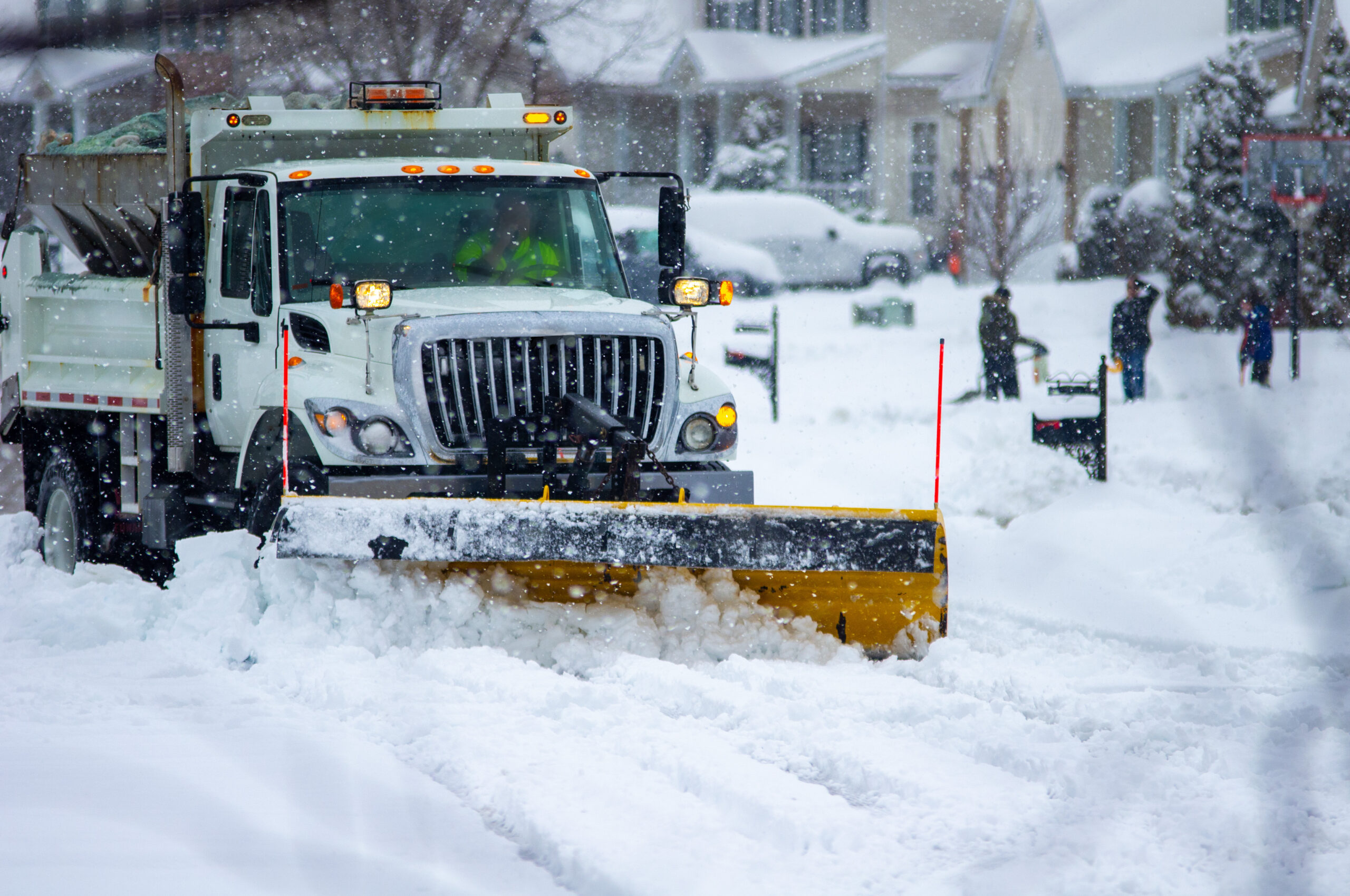 Front view of city services snow plow truck with yellow blade cleaning roads after winter storm with kids playing in the background